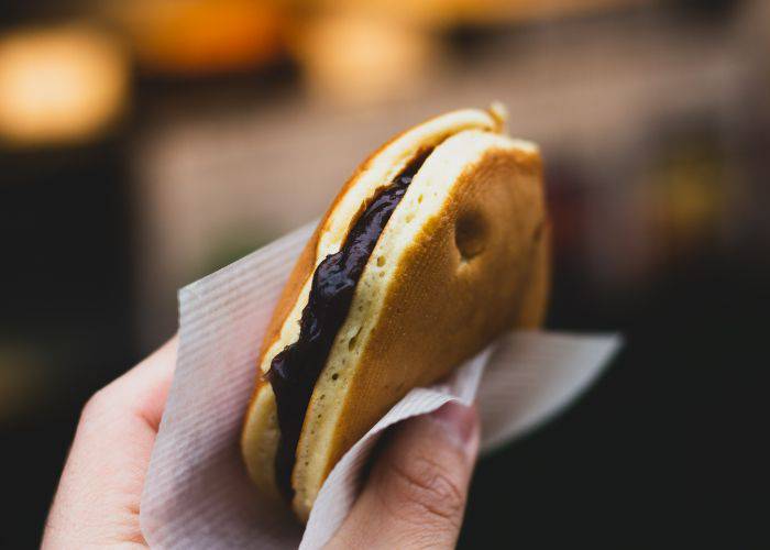 A hand holding a fresh red bean pancake within a napkin. Red beans ooze out of the sponge.